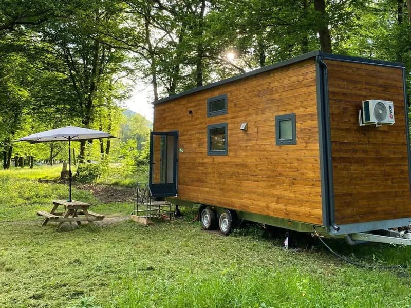 Tiny house Sharewood en bois sur sa remorque, avec parasol et table de pique-nique sous les arbres
