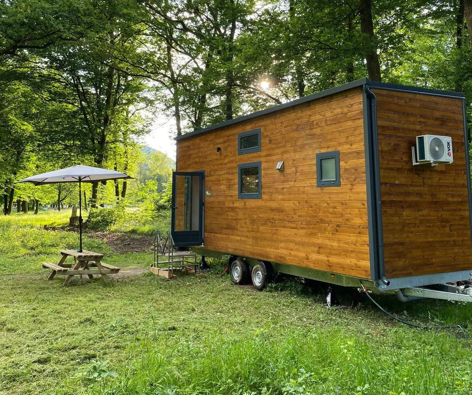 Tiny house Sharewood en bois sur sa remorque, avec parasol et table de pique-nique sous les arbres